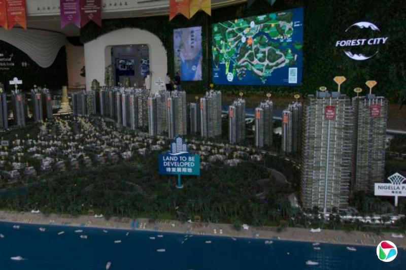 一个城市模型,背景是人造海洋,一块巨大的标牌上写着“待开发用地”,矗立在人造的树木绿地中,标牌后面是模型高楼,模型后面的墙上有一个大萤幕,上面显示着地图和“森林城巿”的标志。